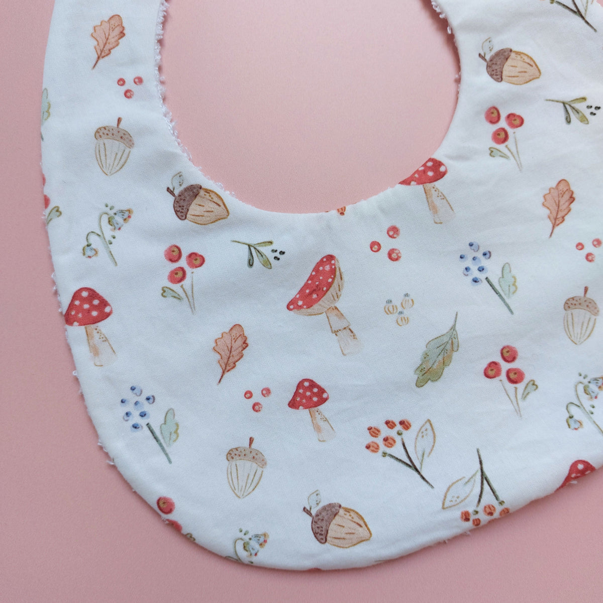 White round shaped baby bib with autumnal pattern of mushrooms, acorns, leaves and autumn flowers. 