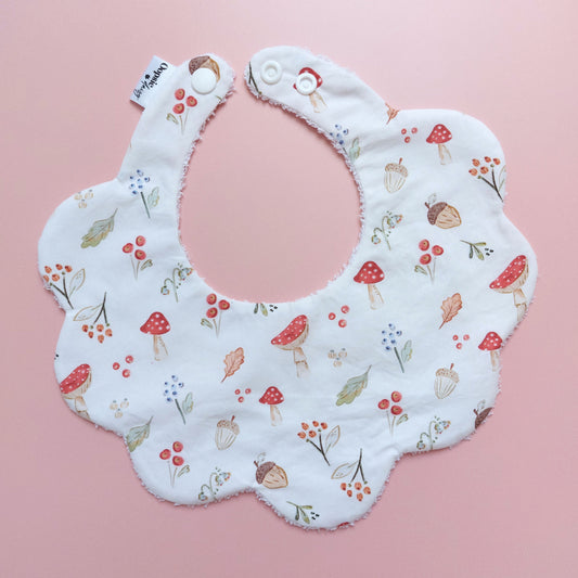 Scallop edged baby bib in white with autumnal pattern. Featuring mushrooms, acorns, leaves and autumnal flowers. 