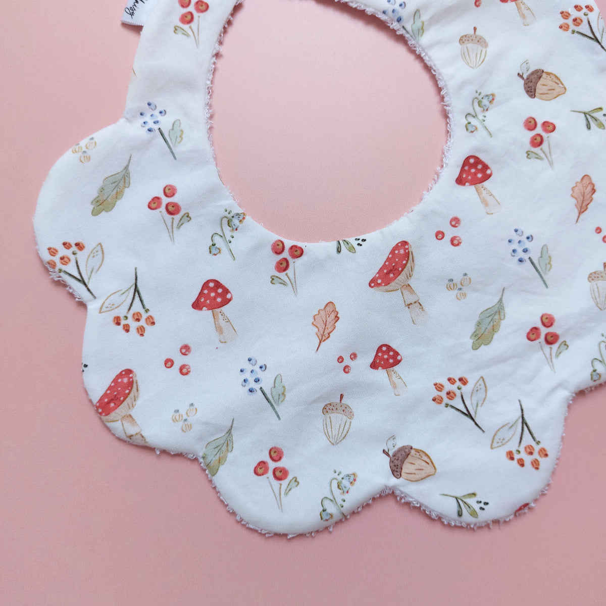 Scallop edged baby bib in white with autumnal pattern. Featuring mushrooms, acorns, leaves and autumnal flowers. 