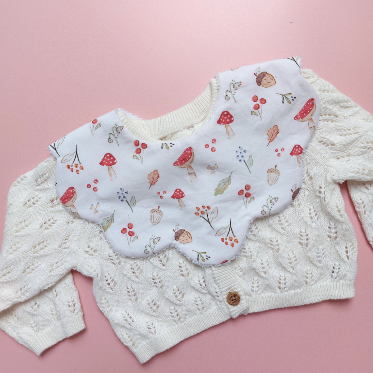 Scallop edged baby bib in white with autumnal pattern. Featuring mushrooms, acorns, leaves and autumnal flowers. 