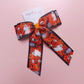 A Halloween-themed grosgrain ribbon bow with a orange, white and black pattern featuring ghosts, skulls, pumpkins, and tombstones.