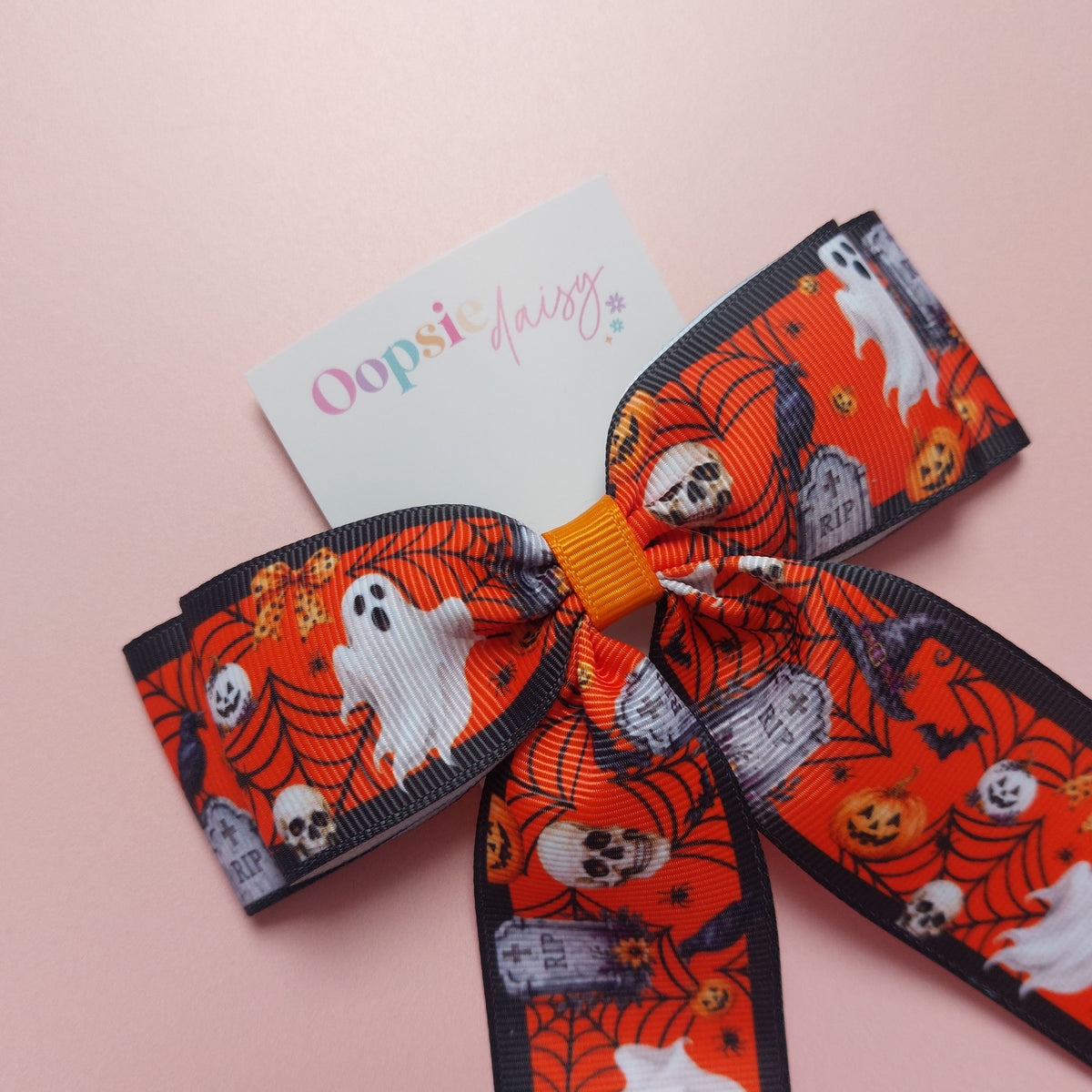 A Halloween-themed grosgrain ribbon bow with a orange, white and black pattern featuring ghosts, skulls, pumpkins, and tombstones.