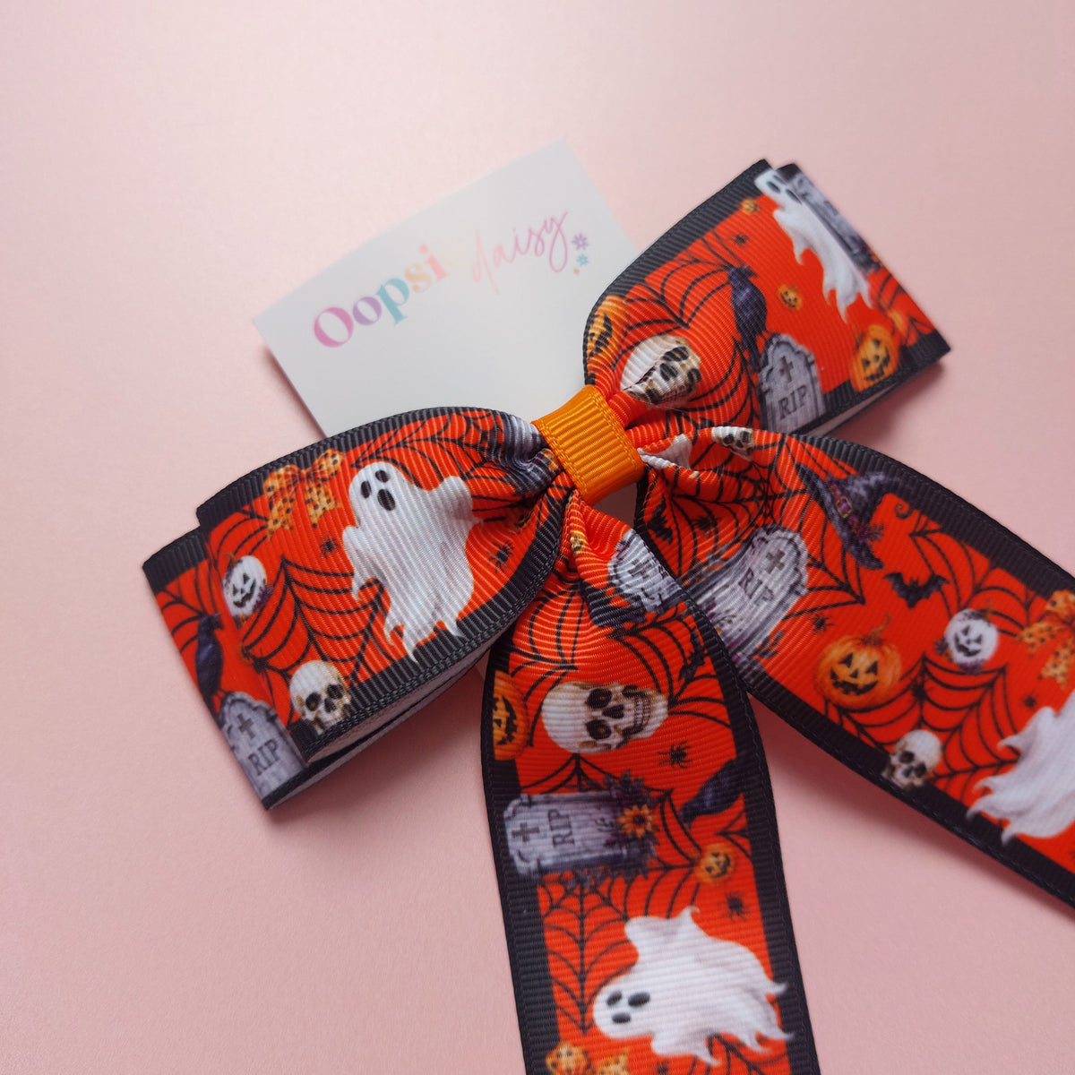 A Halloween-themed grosgrain ribbon bow with a orange, white and black pattern featuring ghosts, skulls, pumpkins, and tombstones.