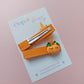 A pair of orange ribbon hair clips with pumpkin embellishment, displayed on a white card from the brand Oops Daisy against a soft pink background.