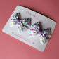 Pair of bow hair clips with bold purple rainbow pattern. 
