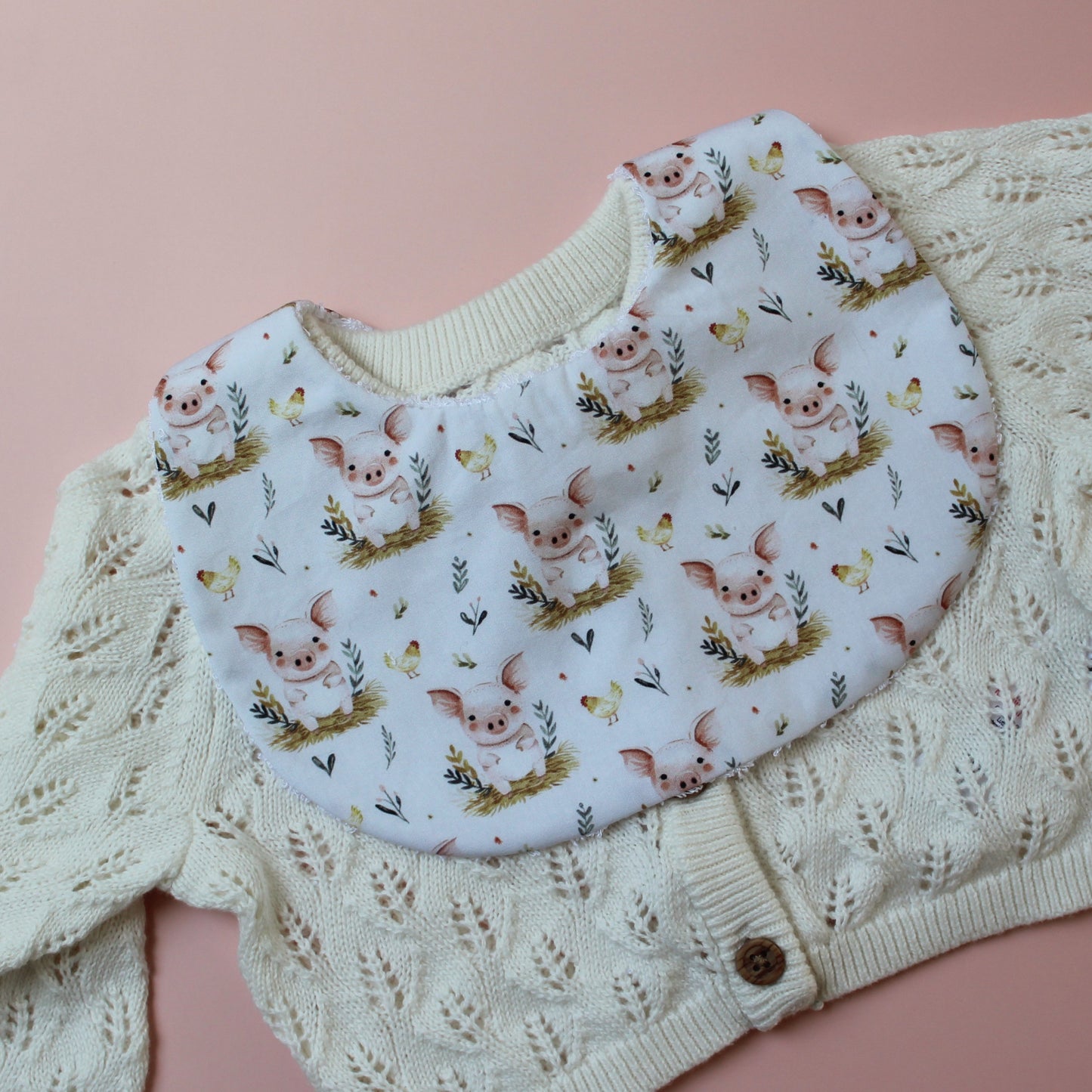 Baby bib with cute pig pattern. White background with pink pigs on bed of barley.