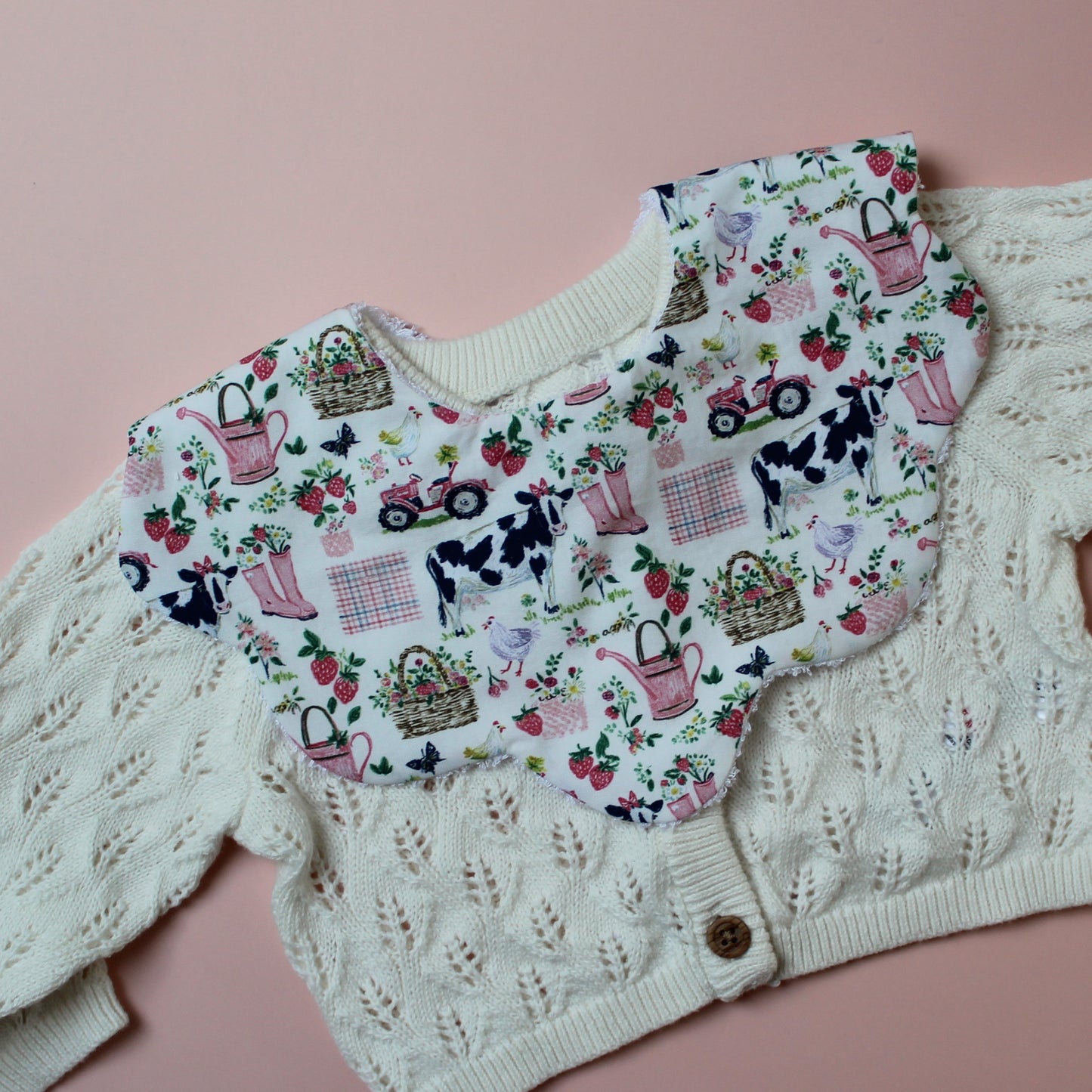 Scallop baby bib with girls farm themed pattern. Cream background with cows, pink watering cans, tractors and strawberries.