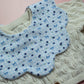 A close-up of a baby bib with a scalloped edge, featuring a blue and white gingham pattern with small blue bows and cherries. The bib is laid flat on top of a cream-colored cable-knit cardigan against a soft pink background.