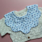 A baby bib with light blue gingham fabric with small blue bows and blueberries layered on an off-white cardigan, laid flat on a pink background.