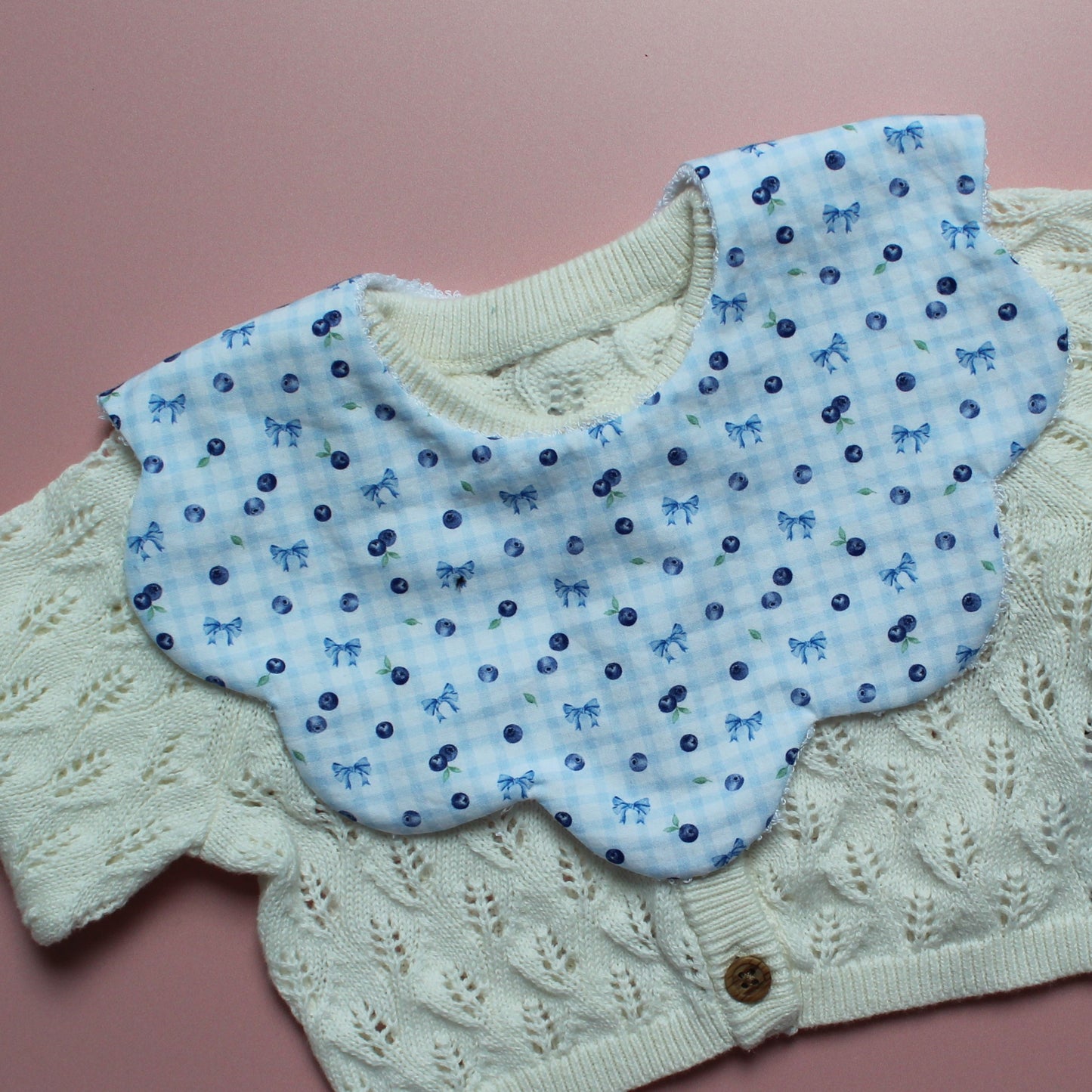 A baby bib with light blue gingham fabric with small blue bows and blueberries layered on an off-white cardigan, laid flat on a pink background.