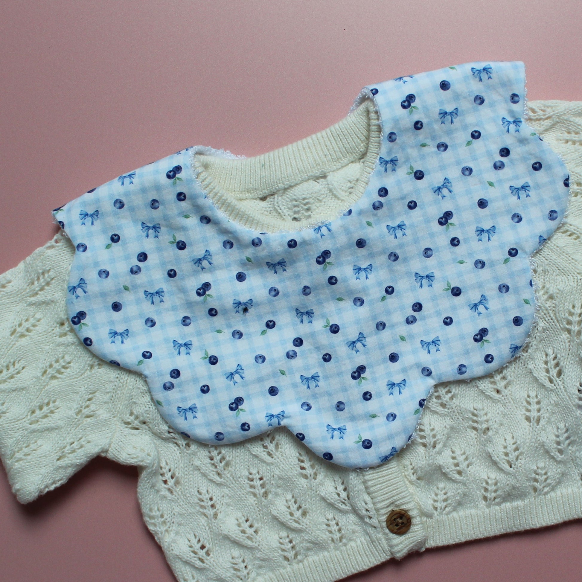 A baby bib with light blue gingham fabric with small blue bows and blueberries layered on an off-white cardigan, laid flat on a pink background.