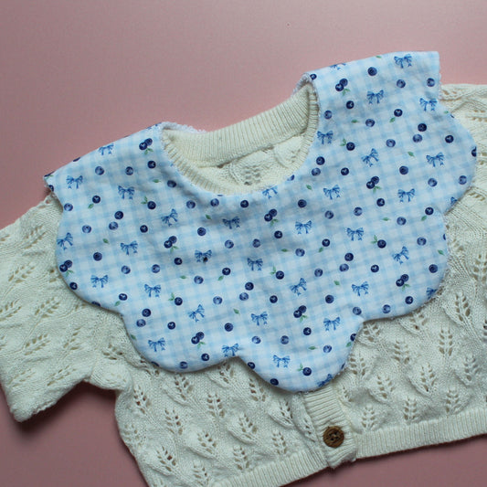 A baby bib with light blue gingham fabric with small blue bows and blueberries layered on an off-white cardigan, laid flat on a pink background.