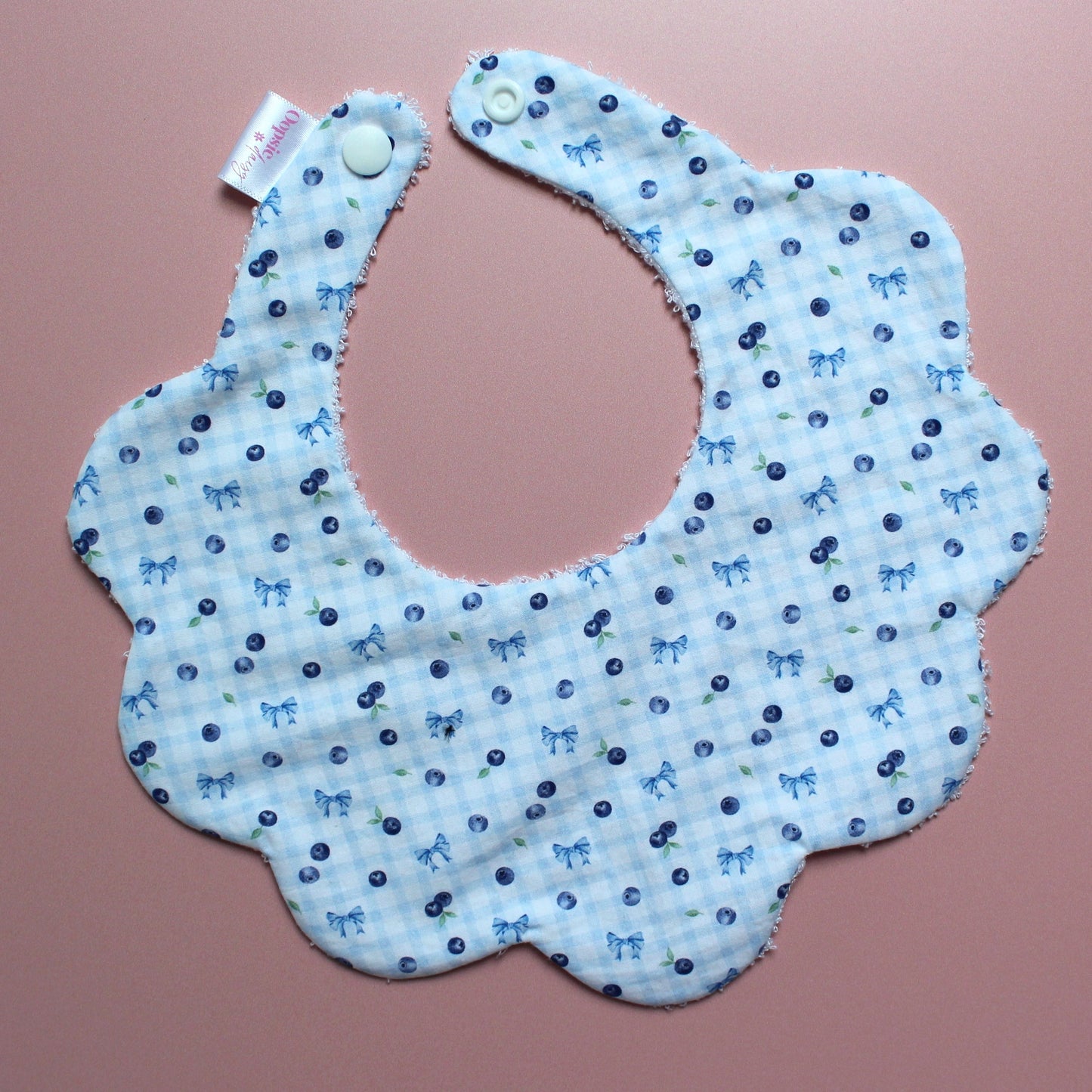 A scalloped fabric bib with a light blue and white gingham pattern, decorated with small blue bows and blueberries, displayed on a pink background.