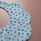 A close-up of a scalloped baby bib made from light blue and white gingham fabric, decorated with a pattern of blueberries and small bows, set against a solid dusty pink background.