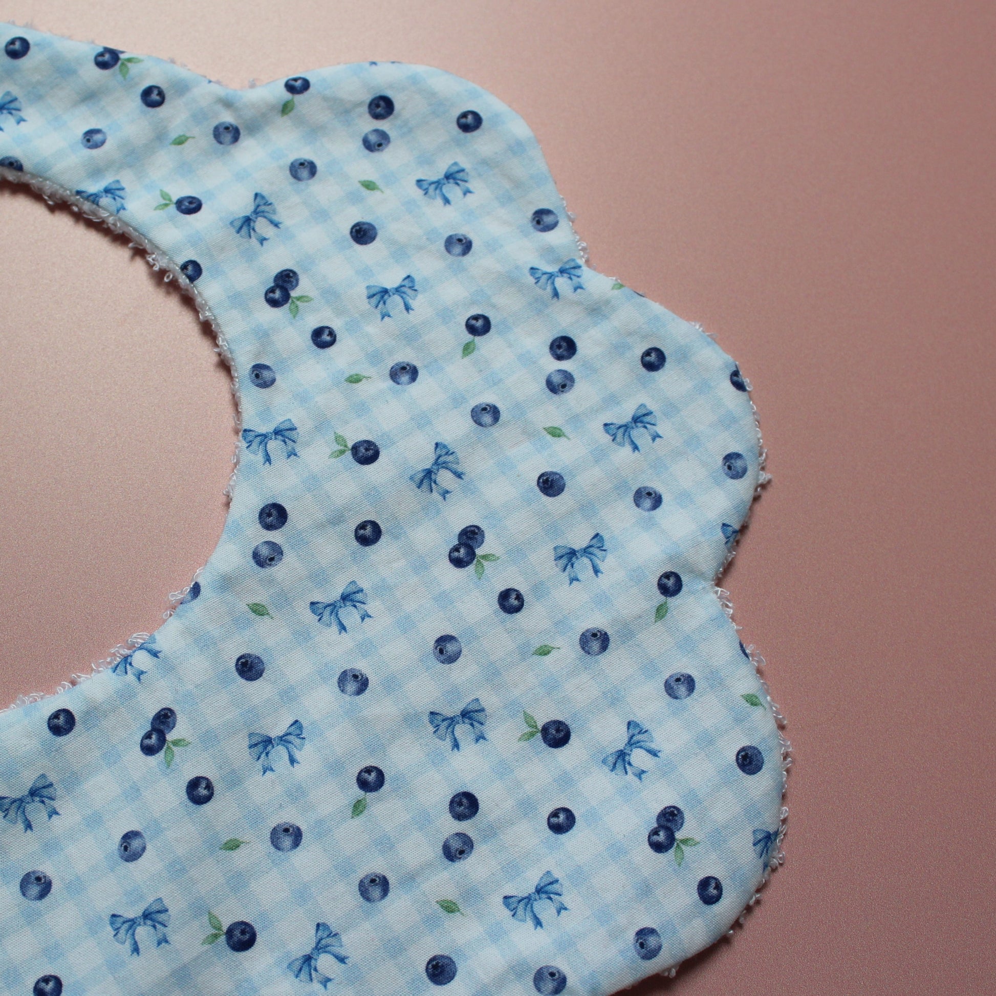 A close-up of a scalloped baby bib made from light blue and white gingham fabric, decorated with a pattern of blueberries and small bows, set against a solid dusty pink background.