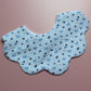 A light blue and white gingham bib with a blue bow and blueberry pattern, laid flat on a dusty pink background.