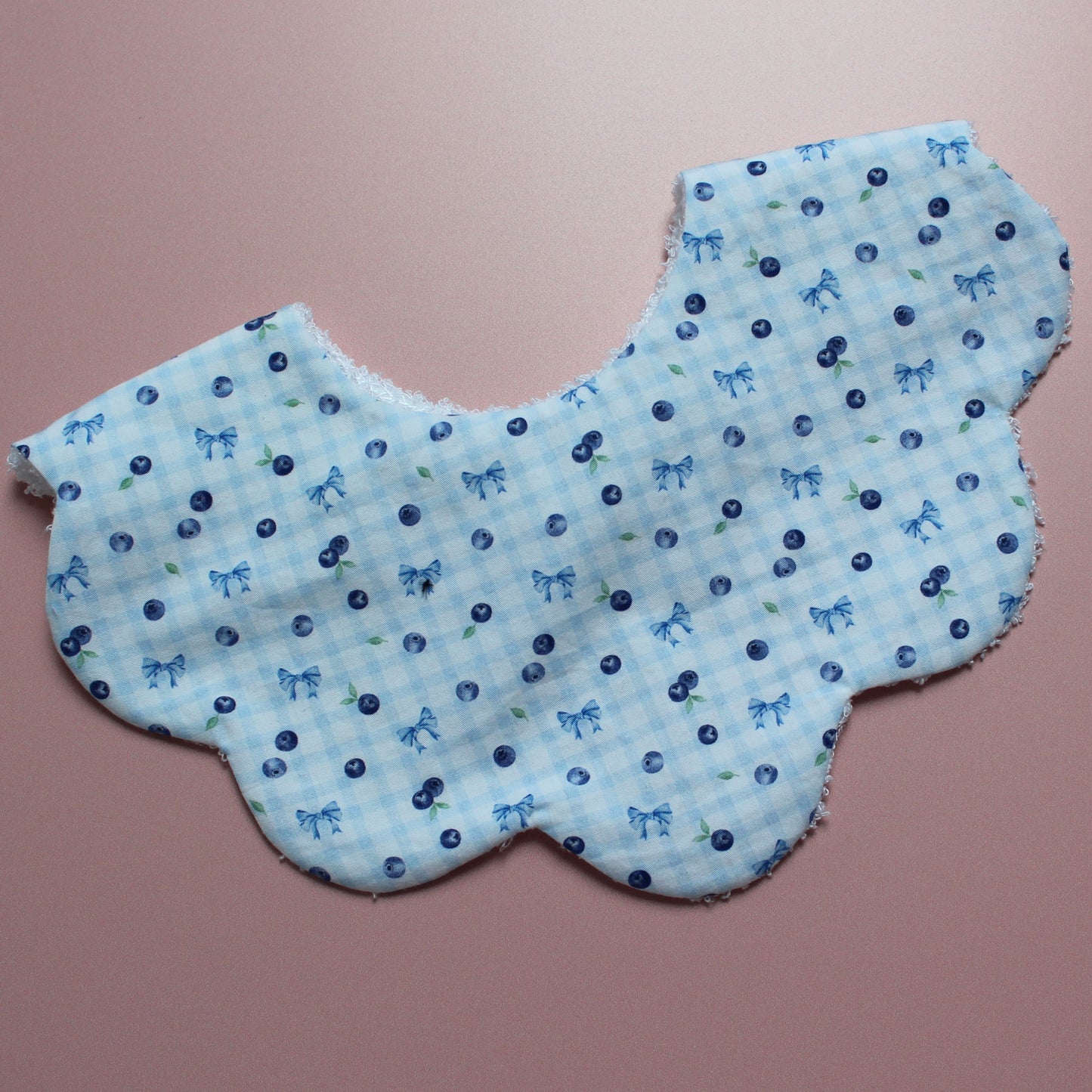 A light blue and white gingham bib with a blue bow and blueberry pattern, laid flat on a dusty pink background.