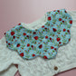 A close-up of a baby bib featuring a blue fabric with a strawberry and white flower print, layered over an off-white cable-knit cardigan on a pink background.