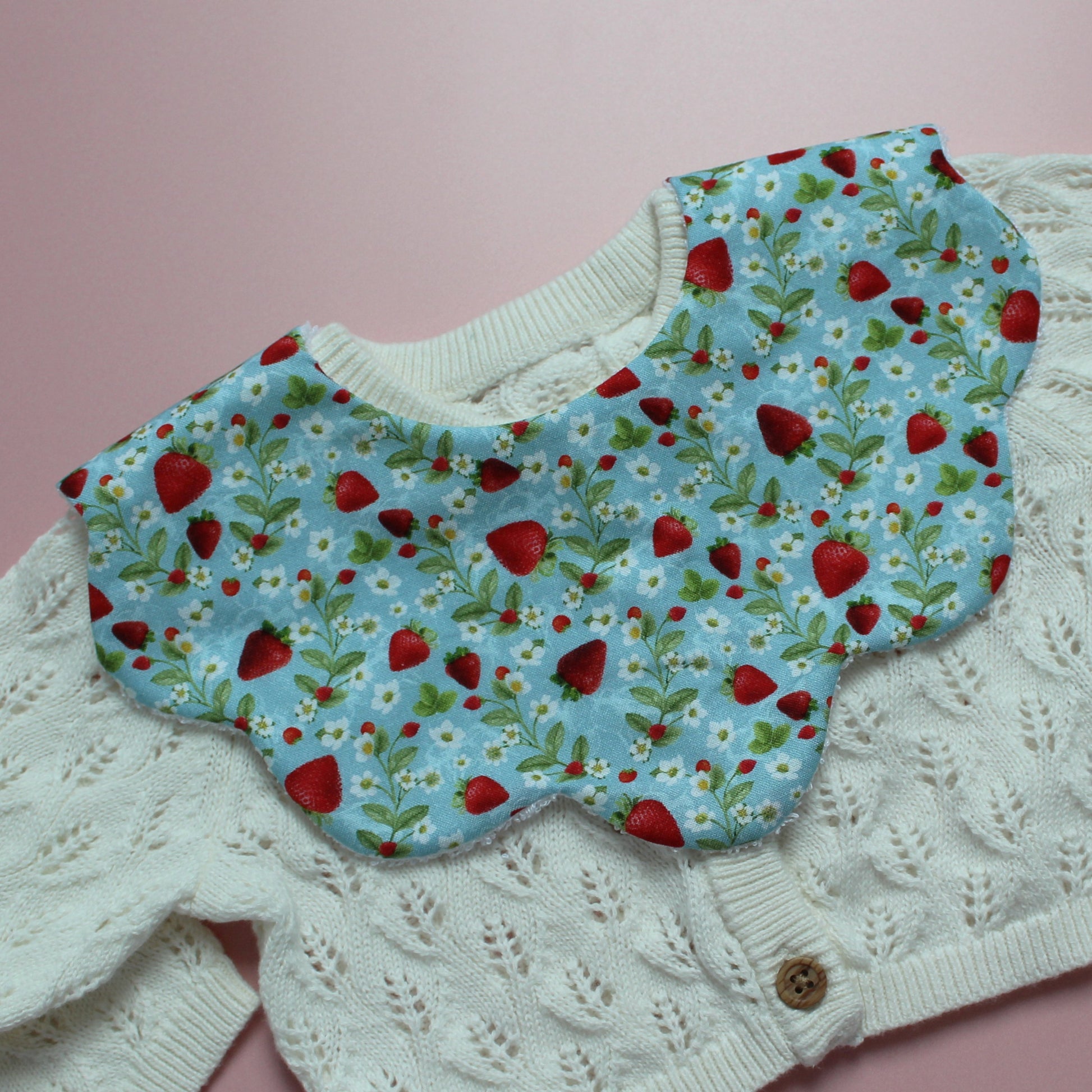 A close-up of a baby bib featuring a blue fabric with a strawberry and white flower print, layered over an off-white cable-knit cardigan on a pink background.