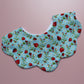 A scalloped fabric bib with a blue background, featuring a repeating pattern of red strawberries, green leaves, and small white flowers, laid flat on a solid pink background.
