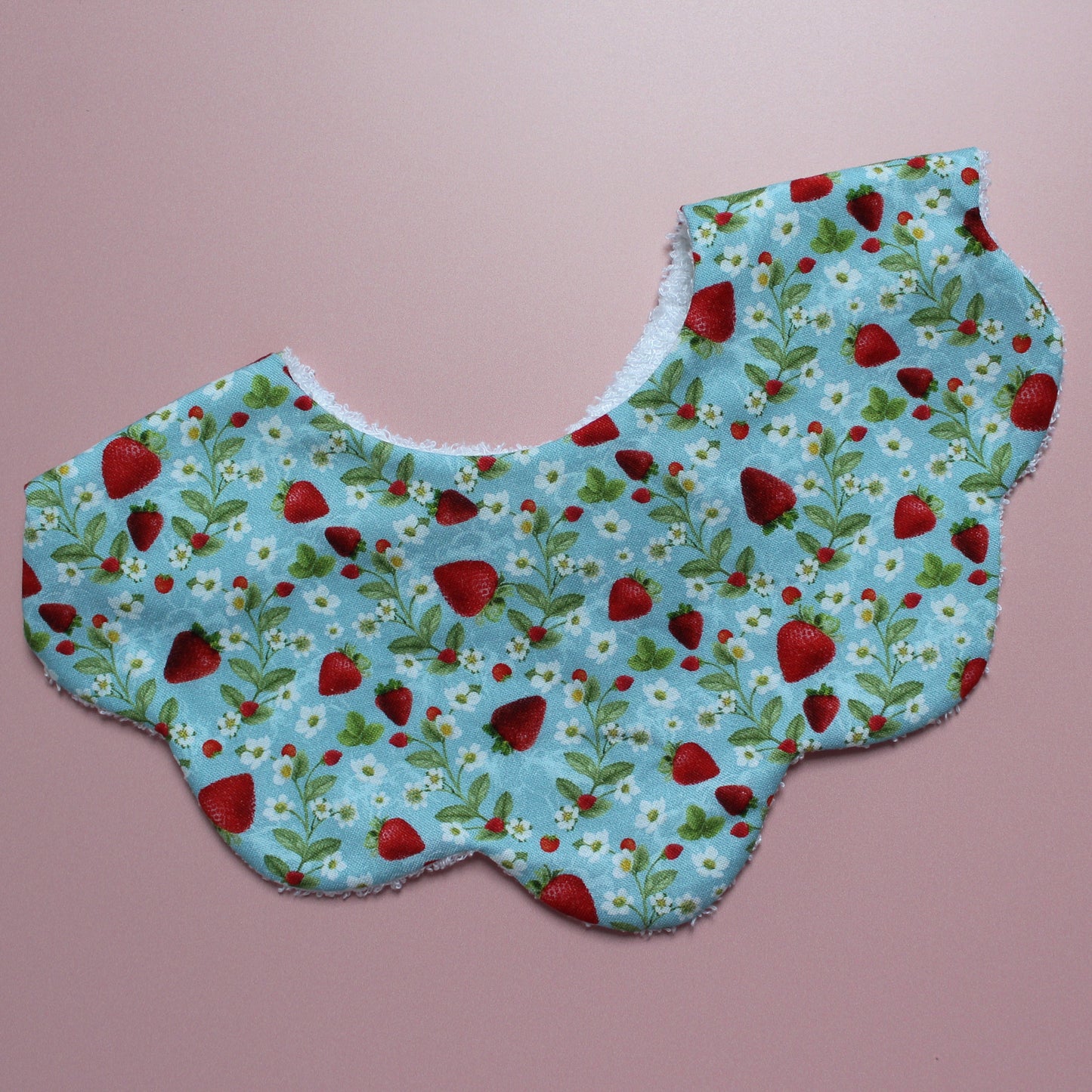 A scalloped fabric bib with a blue background, featuring a repeating pattern of red strawberries, green leaves, and small white flowers, laid flat on a solid pink background.