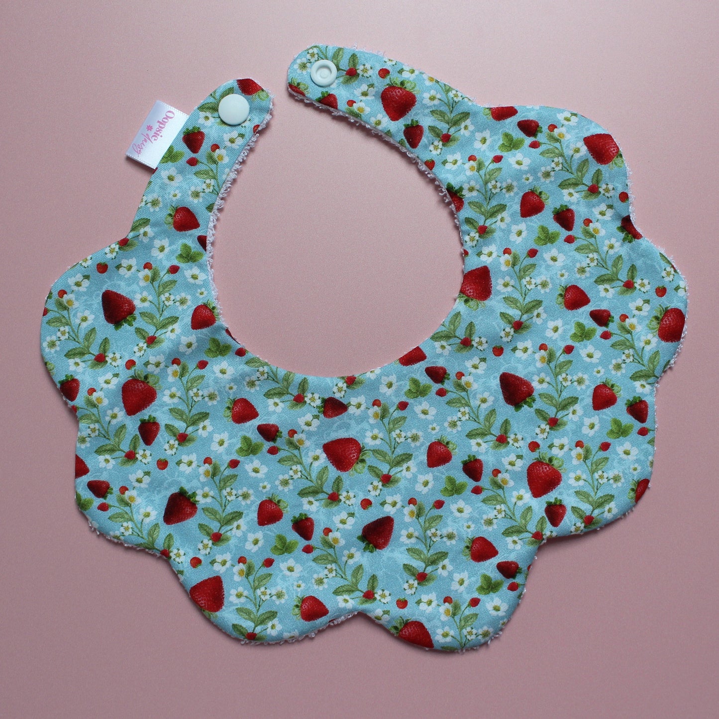 A scalloped fabric bib with a blue background, printed with a red strawberry and white floral pattern, featuring white snap buttons on the neck strap, laid flat on a pink background.