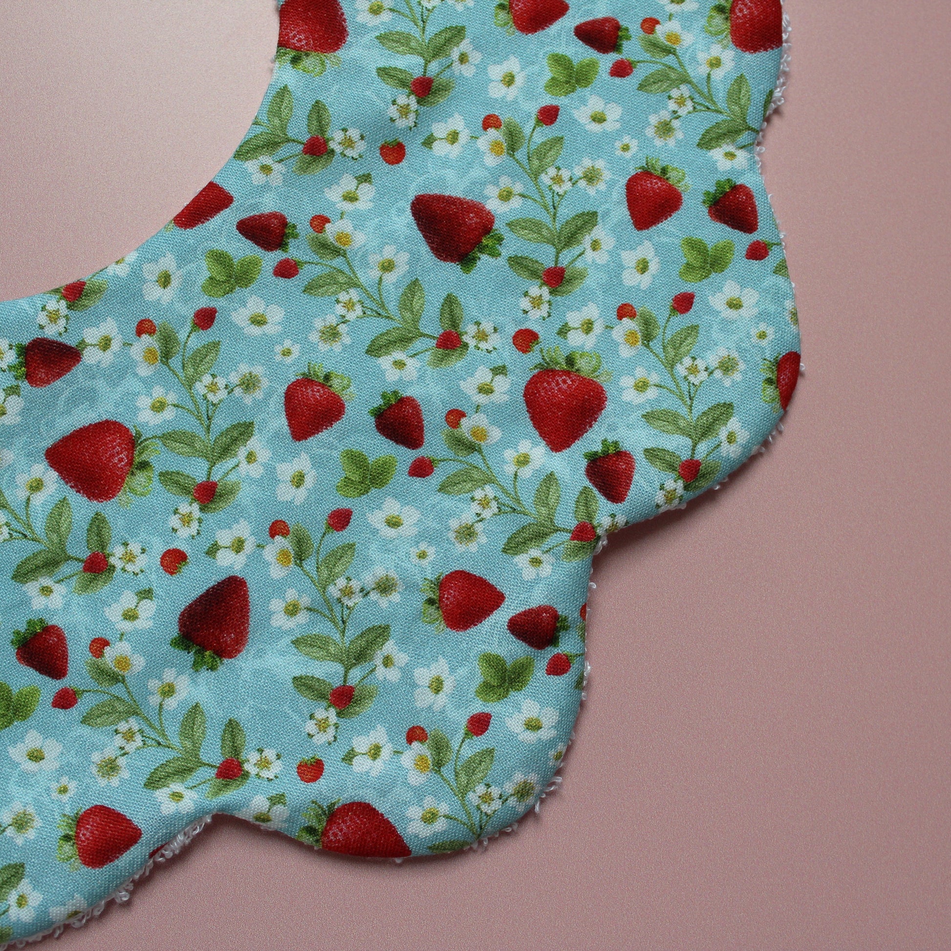 A close-up of a fabric baby bib featuring a blue background with a strawberry and white flower print, laid on a pink background.