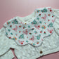 A close-up of a baby bib featuring a white bib with a pink chicken and floral print layered over an off-white cable-knit cardigan, set against a pink background.