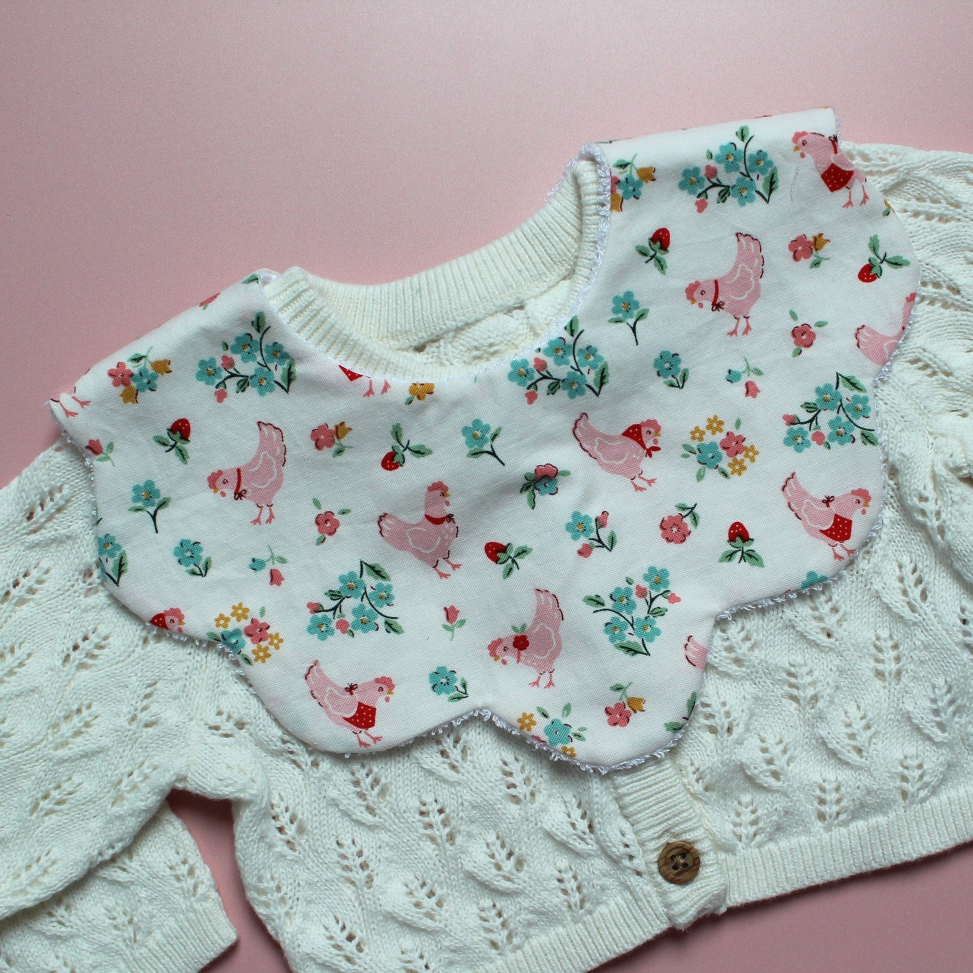 A close-up of a baby bib featuring a white bib with a pink chicken and floral print layered over an off-white cable-knit cardigan, set against a pink background.