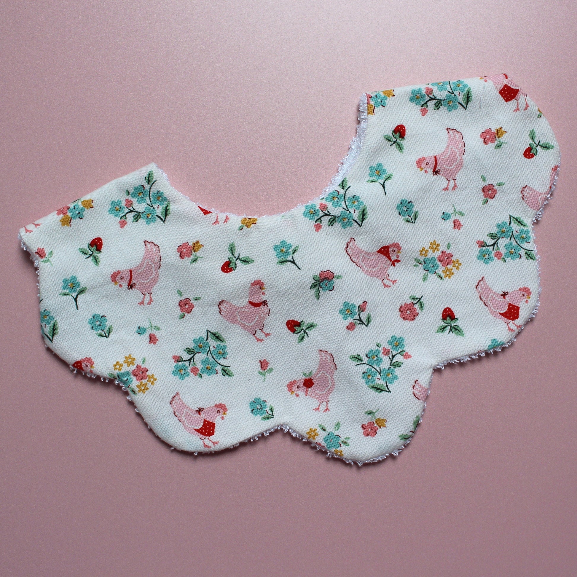 A scalloped-edge fabric bib laid flat on a pink background. The white fabric features a colourful print of pink chickens, red strawberries, and green and blue flowers.