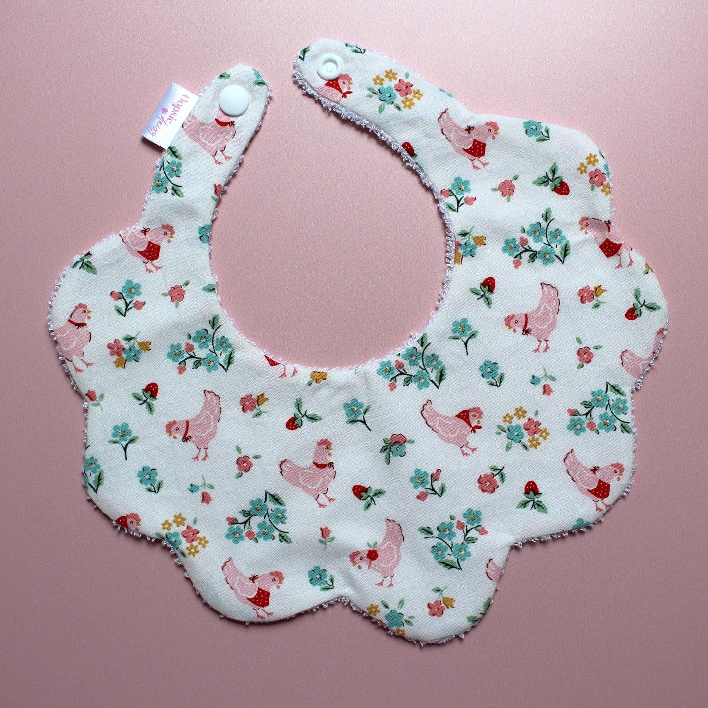 A scalloped baby bib with a white background and a vintage-style print of pink chickens and green and red flowers, laid flat on a pink background.