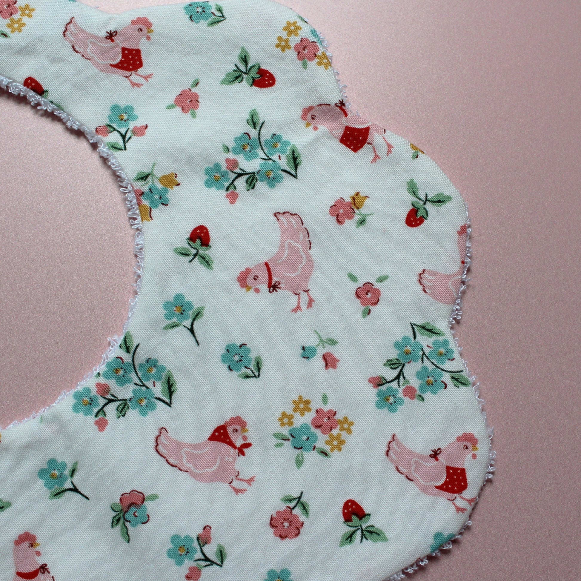 A close-up of a scalloped fabric bib featuring a white background with a pink, blue, and red floral and chicken pattern, laid flat on a pink background.
