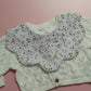 A close-up of a baby bib featuring a pale pink floral bib with white flowers and green centers, layered over a cream-colored cable-knit cardigan.