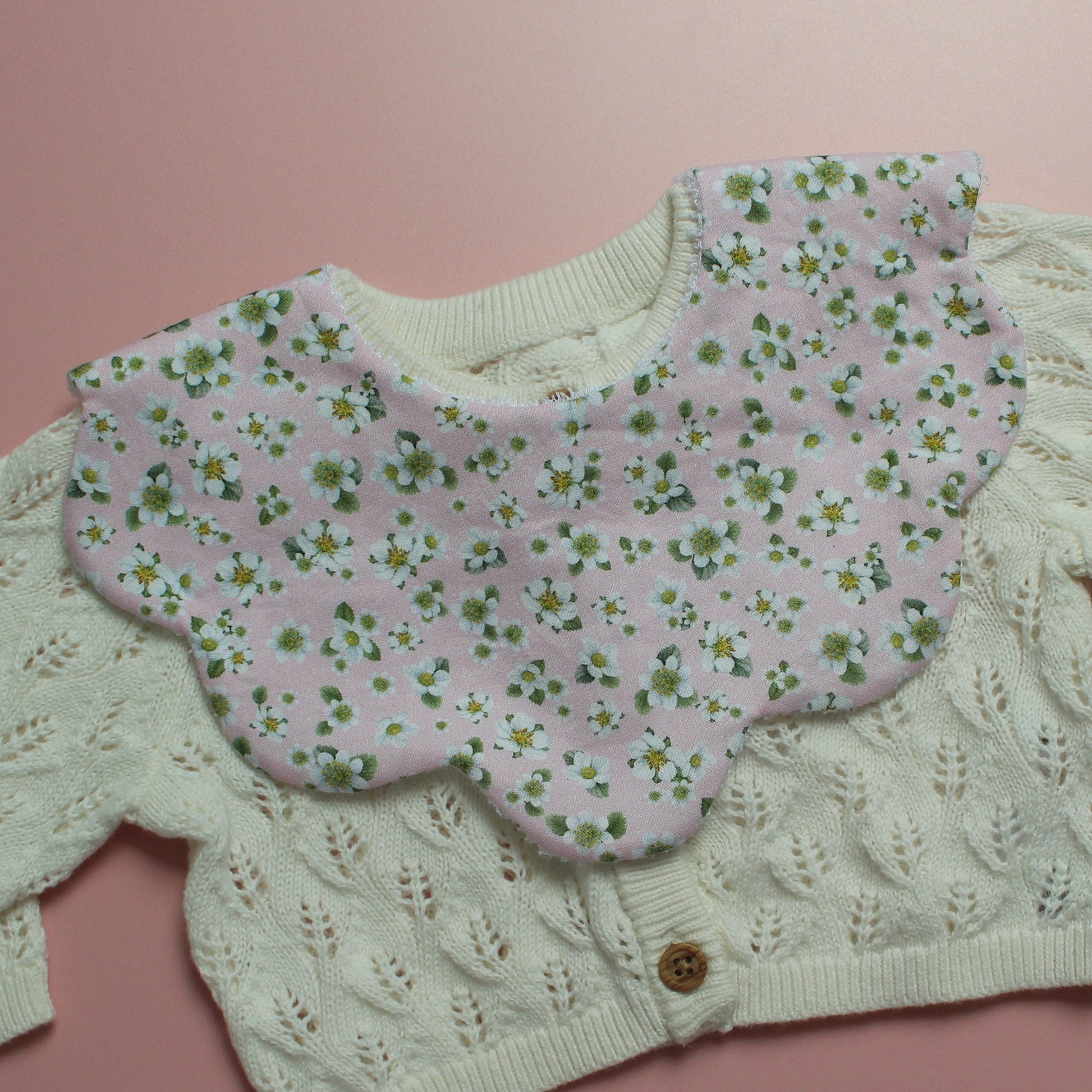 A close-up of a baby bib featuring a pale pink floral bib with white flowers and green centers, layered over a cream-colored cable-knit cardigan.
