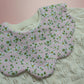 A close-up of a pink fabric bib with a white and green floral pattern, laid over a cream-colored cable-knit cardigan.