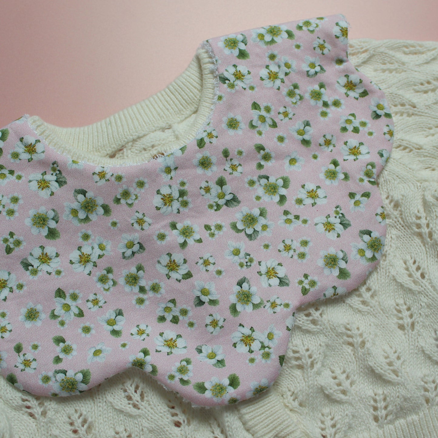 A close-up of a pink fabric bib with a white and green floral pattern, laid over a cream-colored cable-knit cardigan.