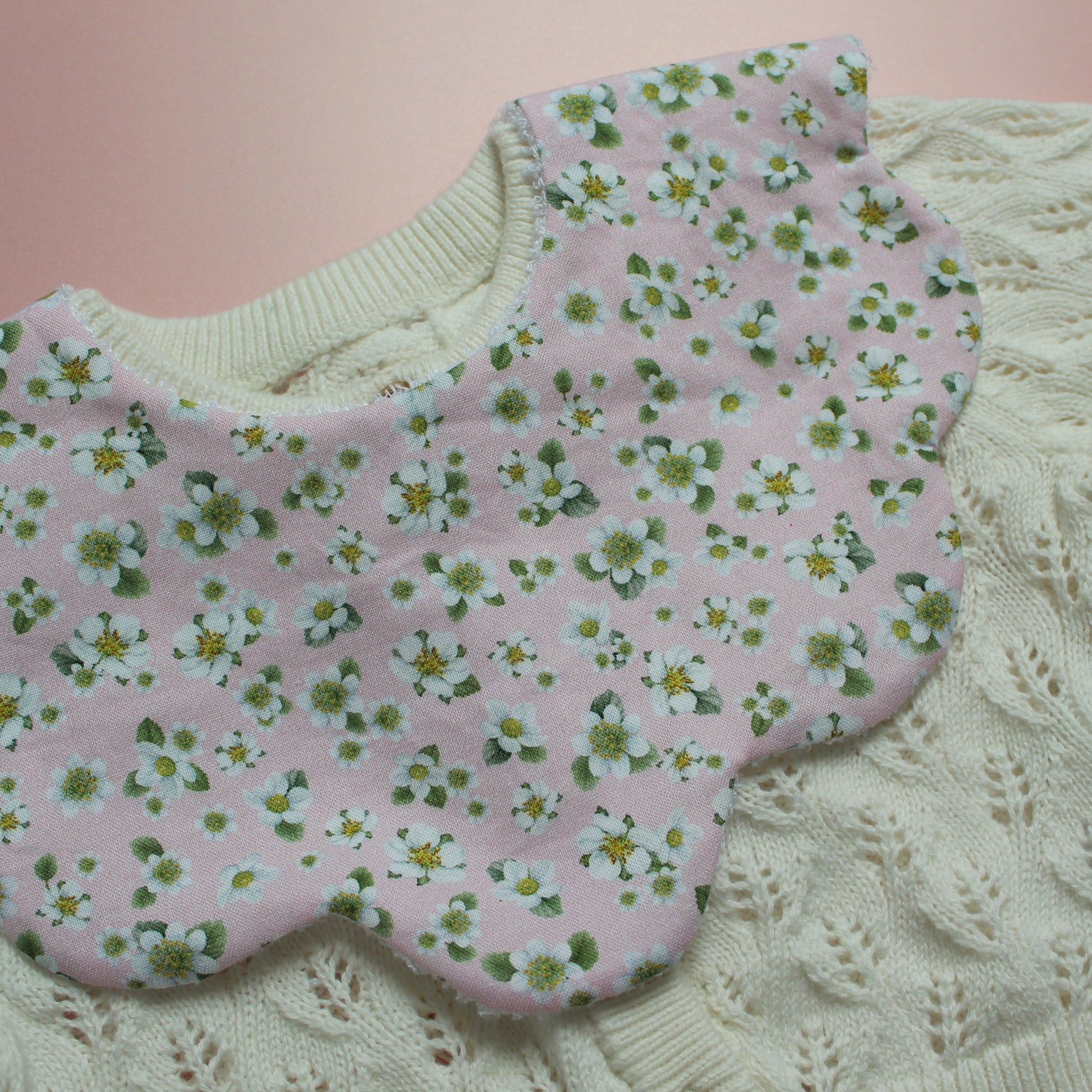 A close-up of a pink fabric bib with a white and green floral pattern, laid over a cream-colored cable-knit cardigan.