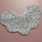 A scalloped-edge fabric bib with a delicate white and green floral print on a light pink background, displayed flat against a solid pink backdrop.