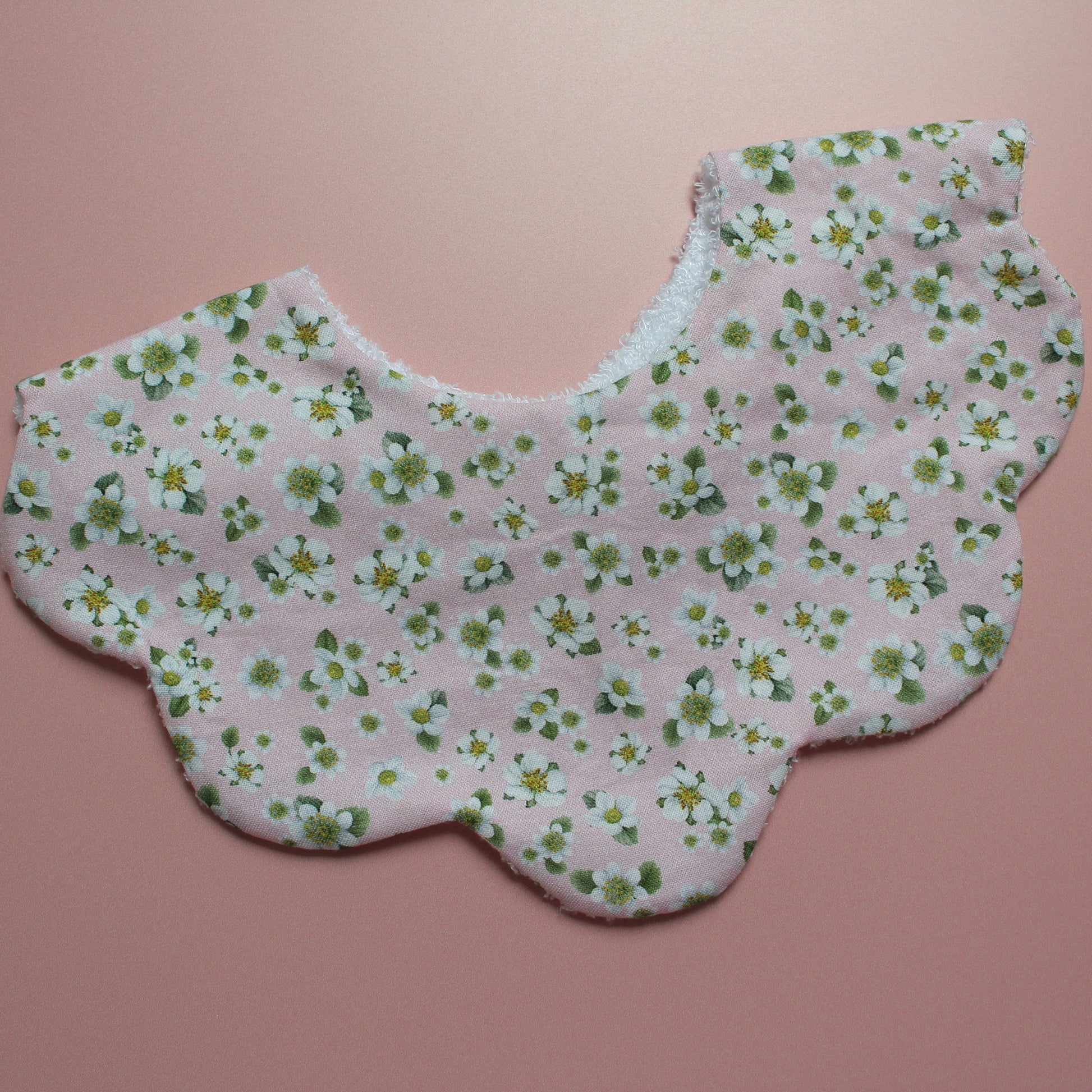 A scalloped-edge fabric bib with a delicate white and green floral print on a light pink background, displayed flat against a solid pink backdrop.