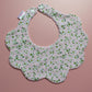 A scalloped baby bib with a light pink background and a green and white floral pattern, featuring snap closures at the top and a small brand tag on the left side, displayed on a solid pink background.