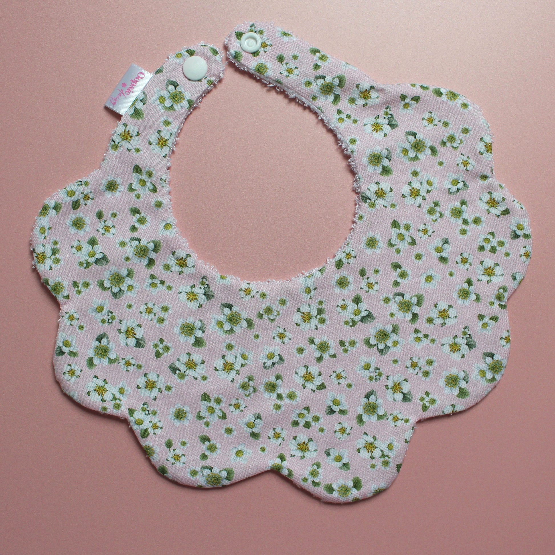 A scalloped baby bib with a light pink background and a green and white floral pattern, featuring snap closures at the top and a small brand tag on the left side, displayed on a solid pink background.
