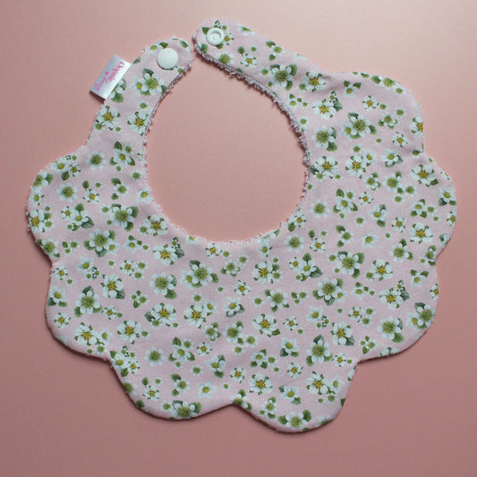 A scalloped baby bib with a light pink background and a green and white floral pattern, featuring snap closures at the top and a small brand tag on the left side, displayed on a solid pink background.