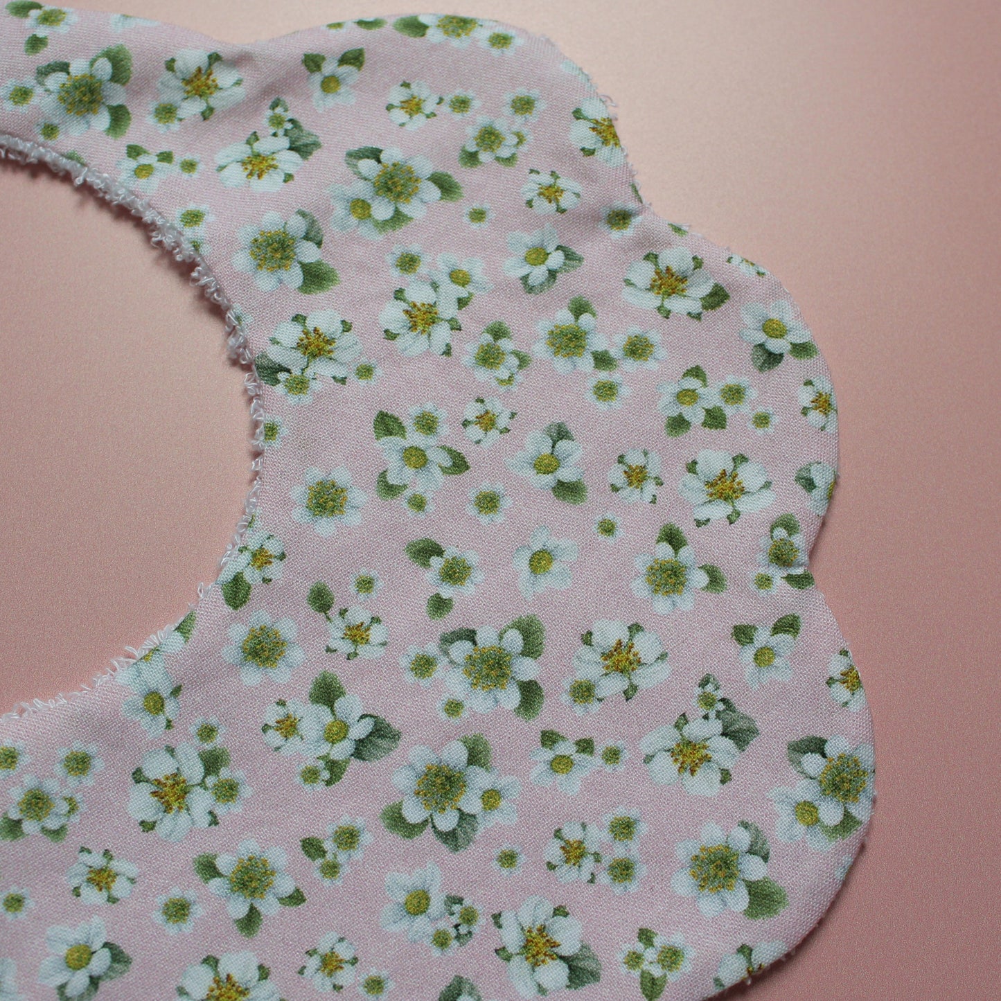 A close-up of a scalloped baby bib made from light pink fabric with a white and green floral print, laid flat on a solid pink background.