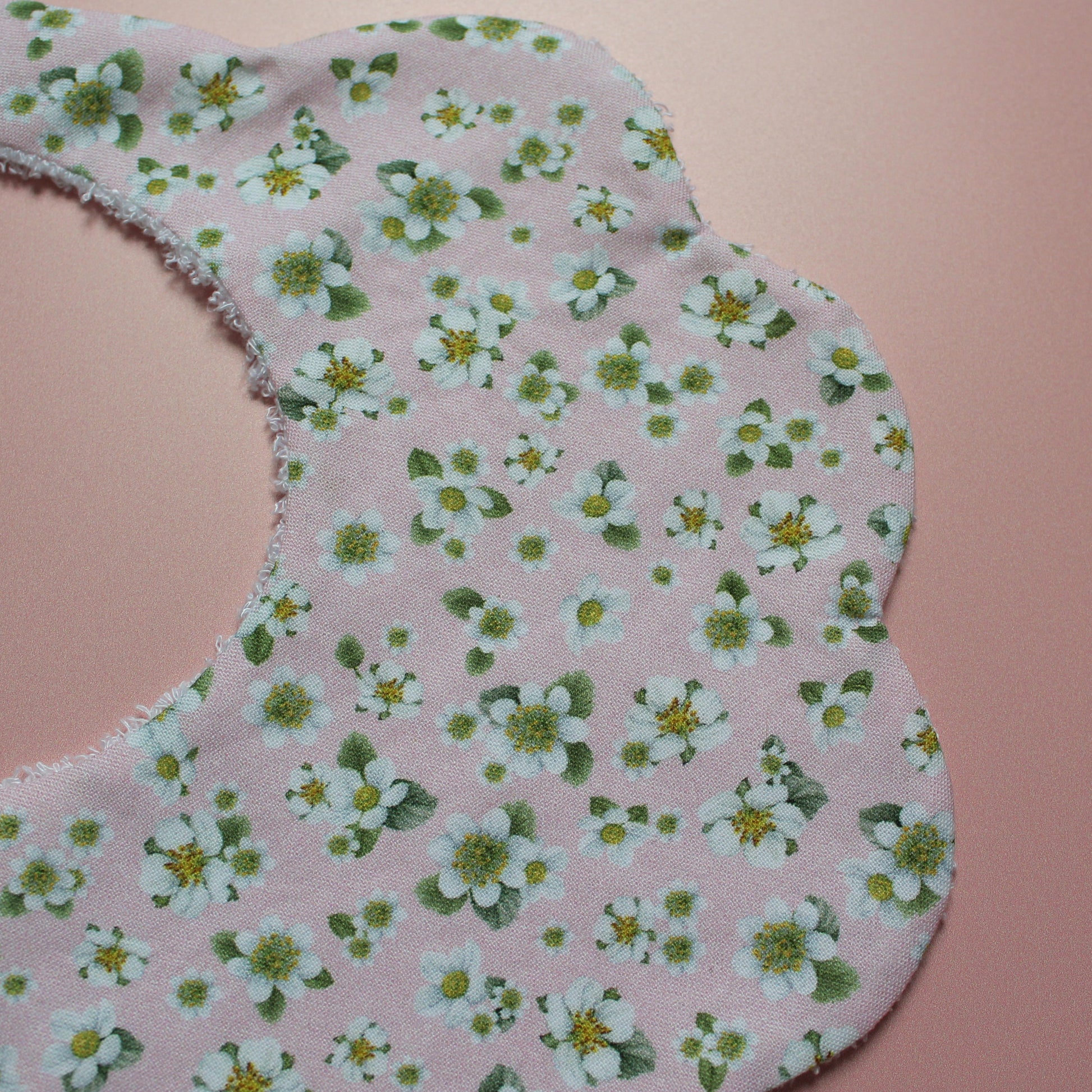A close-up of a scalloped baby bib made from light pink fabric with a white and green floral print, laid flat on a solid pink background.
