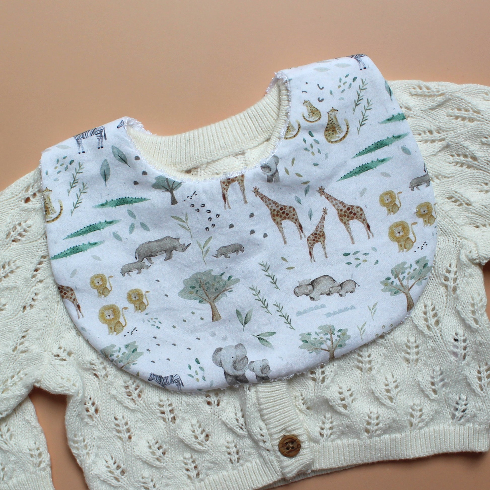 A baby bib with an animal safari print, styled on top of a cream-colored cable-knit cardigan against a solid peach background.