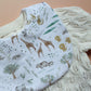 A close-up of a white baby bib with a safari animal print, laid over a cream-colored cable-knit cardigan on a tan background.