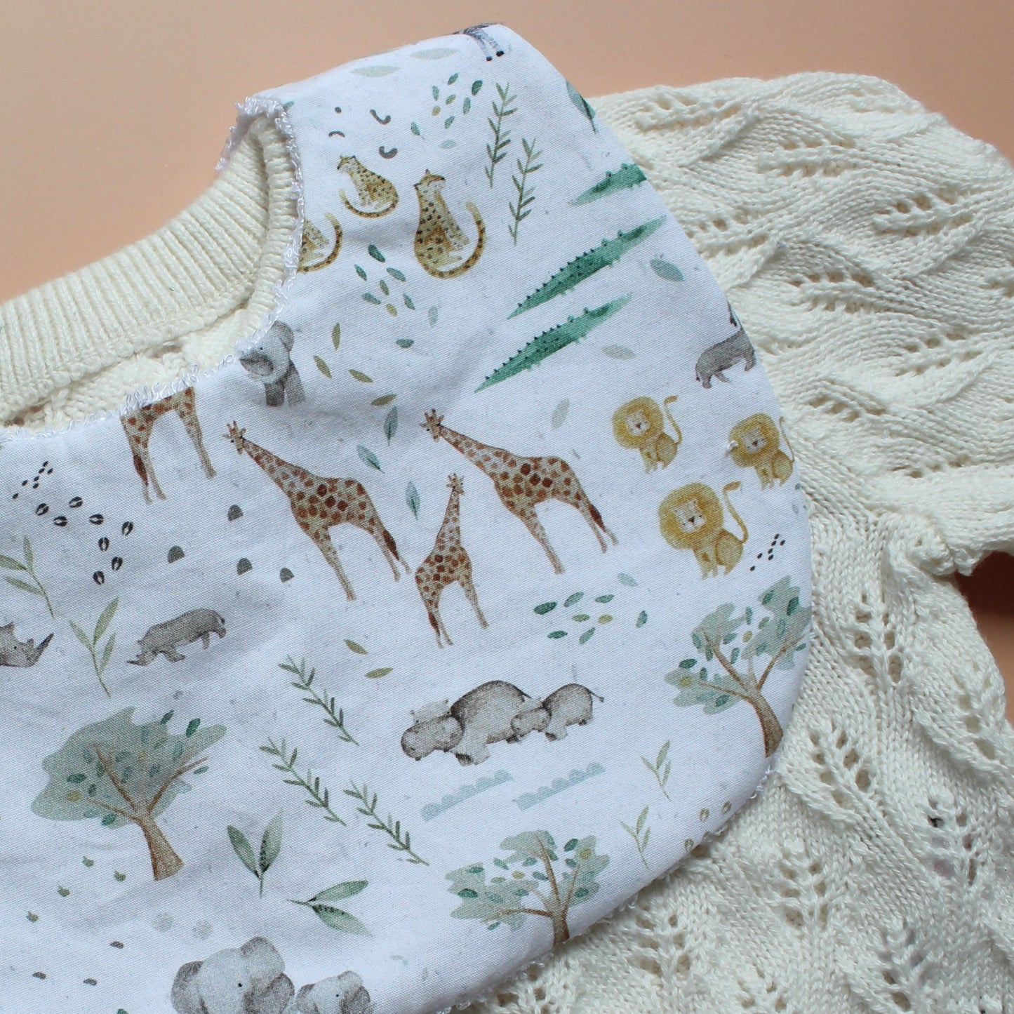 A close-up of a white baby bib with a safari animal print, laid over a cream-colored cable-knit cardigan on a tan background.