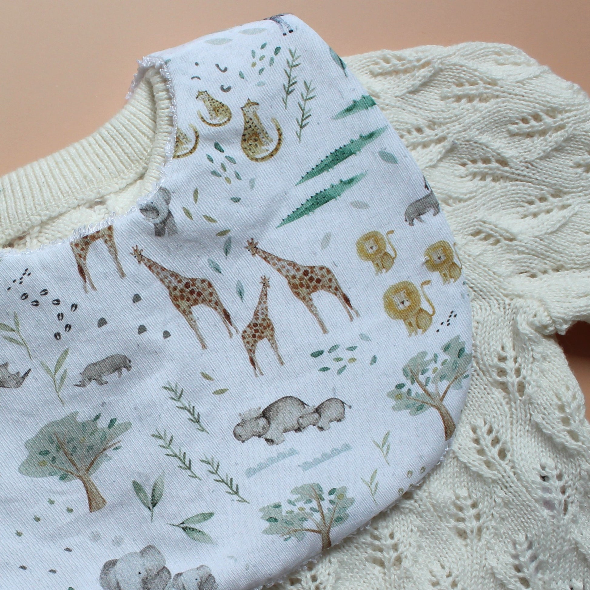 A close-up of a white baby bib with a safari animal print, laid over a cream-colored cable-knit cardigan on a tan background.