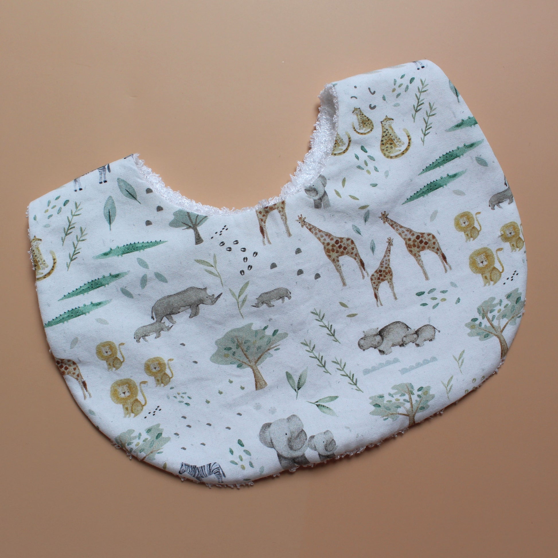 A white fabric bib with a safari animal print featuring giraffes, elephants, hippos, and trees, laid flat on a solid tan background.