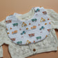 A baby bib with a construction vehicle print, layered over a cream-colored cable-knit cardigan sweater, laid flat on a solid tan background.